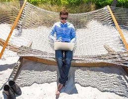 joven que trabaja con una laptop en una hamaca durante las vacaciones en la playa foto