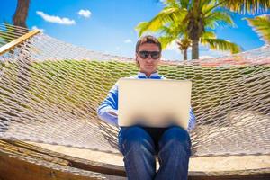 Young man working with laptop in hammock during beach vacation photo