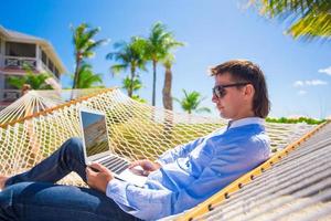 Young man working with laptop in hammock during beach vacation photo