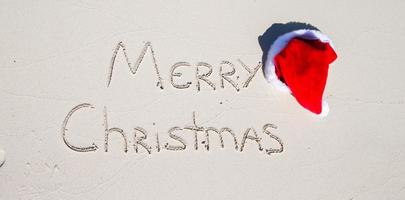 Santa Hat on white sandy beach and Merry Christmas written in the sand photo