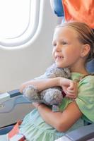 Adorable little girl traveling by an airplane. Kid sitting near aircraft window with her favourite toy photo