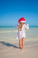 Back view of Little cute girl in red hat santa claus on the beach photo