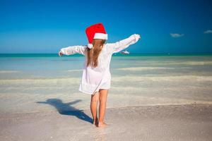 vista trasera de una niña linda con sombrero rojo santa claus en la playa foto