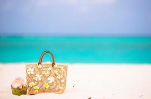 Beach bag with frangipani flowers and coconut on white beach. Closeup beach accessories. photo
