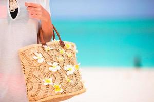 Closeup beautiful bag with frangipani flowers and sunglasses on white beach in female hands photo