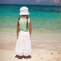 vista trasera de una niña con sombrero mirando el mar en una playa de arena blanca foto