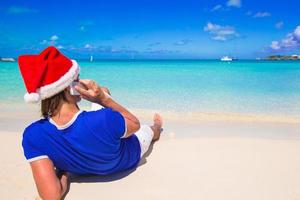 joven con gorro de Papá Noel hablando por teléfono en una playa tropical foto