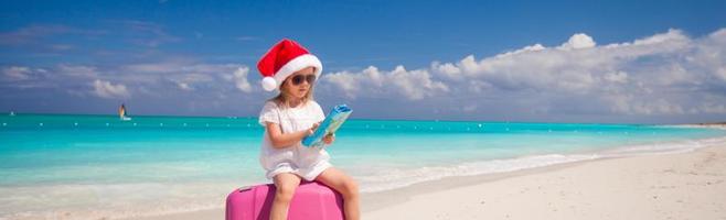 niña pequeña con sombrero de santa sentada en una maleta grande en una playa tropical foto