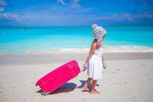Adorable girl in warm winter hat and mittens walking with luggage on beach photo