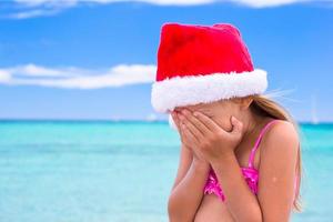 Little adorable girl in red Santa hat at tropical beach photo