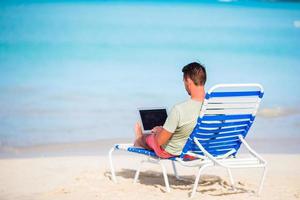 Young man with laptop on tropical caribbean beach. Man sitting on the sunbed with computer and working on the beach photo