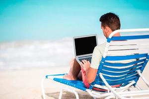 Young man with laptop on tropical caribbean beach. Man sitting on the sunbed with computer and working on the beach photo