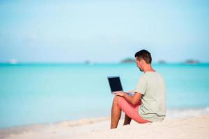 joven sentado en la arena con una computadora portátil en una playa caribeña tropical. hombre con computadora y trabajando en la playa foto