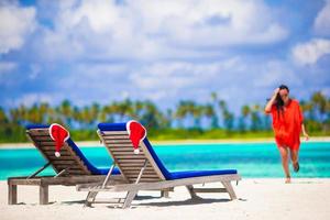 Two loungers with red Santa Hats on tropical white beach with turquoise water. Young woman walking on the beach photo