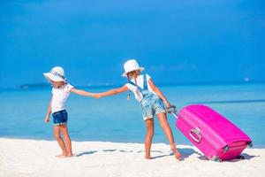 Little tourists girls with big suitcase on tropical white beach. Travel concept. photo