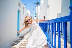 Charming girl in white dress outdoors in old streets an Mykonos. Kid at street of typical greek traditional village with white walls and colorful doors on Mykonos Island, in Greece photo