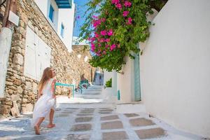 adorable niña con vestido blanco divirtiéndose al aire libre. niño en la calle del típico pueblo tradicional griego con paredes blancas y puertas coloridas en la isla de mykonos, en grecia foto