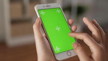 Closeup of hand woman using smartphone with green screen while sitting in living room. Blank digital smartphone in hand girl. Showing content videos blogs tapping on center screen.