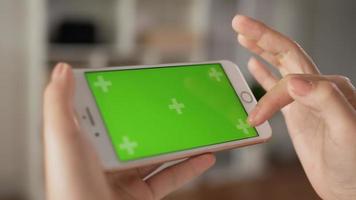 Closeup horizontal of hand woman using smartphone with green screen while sitting in living room. Blank digital smartphone in hand girl. Showing content videos blogs tapping on center screen.