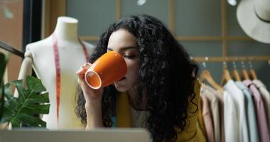 Handheld close up shot, Young beautiful woman fashion designer working in shop drinking beverage in mug while looking on laptop computer, Startup small business entrepreneur concept video