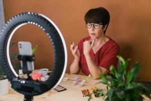 Middle-aged woman talking on cosmetics with makeup eye shadows and blush palette and brushes while recording her video. Mature female making video for her blog on cosmetics photo