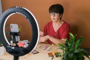 Middle-aged woman talking on cosmetics with makeup eye shadows and blush palette and brushes while recording her video. Mature female making video for her blog on cosmetics photo