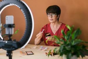 Middle-aged woman talking on cosmetics with makeup eye shadows and blush palette and brushes while recording her video. Mature female making video for her blog on cosmetics photo