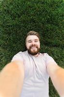 young man with beard and fashion hair style lying on grass taking selfie - holding smart phone or tablet and looking at camera. photo