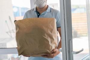 Deliver man wearing face mask in blue uniform handling bag of food give to customer in front of house. Postman and express grocery delivery service photo