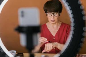 Middle-aged woman talking on cosmetics with makeup eye shadows and blush palette and brushes while recording her video. Mature female making video for her blog on cosmetics photo