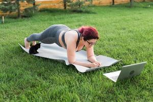 mujer joven de talla grande con top deportivo y polainas de pie en un tablón sobre una alfombra de yoga pasando tiempo en la hierba verde en el patio. gatito negro camina a su alrededor. concepto de bienestar y fitness foto