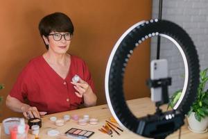 Middle-aged woman talking on cosmetics with makeup eye shadows and blush palette and brushes while recording her video. Mature female making video for her blog on cosmetics photo