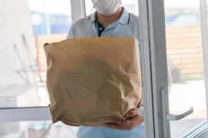 Deliver man wearing face mask in blue uniform handling bag of food give to customer in front of house. Postman and express grocery delivery service photo
