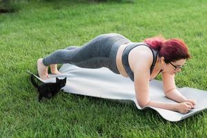 mujer joven de talla grande con top deportivo y polainas de pie en un tablón sobre una alfombra de yoga pasando tiempo en la hierba verde en el patio. gatito negro camina a su alrededor. concepto de bienestar y fitness foto