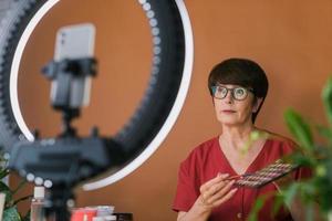 Middle-aged woman talking on cosmetics with makeup eye shadows and blush palette and brushes while recording her video. Mature female making video for her blog on cosmetics photo