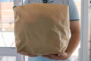 Deliver man wearing face mask in blue uniform handling bag of food give to customer in front of house. Postman and express grocery delivery service photo