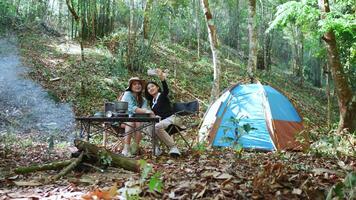 handhållen skott, främre se ung asiatisk Söt kvinna och henne flickvän Sammanträde på främre av tält, använda sig av mobil telefon ta Foto under camping i skog med lycka tillsammans video