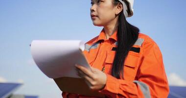 Handheld close up shot, Asian Young engineer woman wearing protection uniform and white helmet standing at front of solar panel to checking operation in paperwork while working in solar farm video