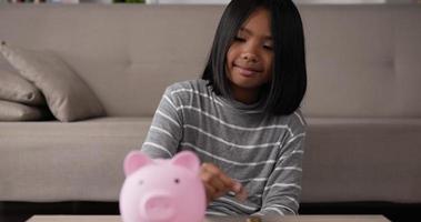 primer plano de una chica asiática feliz poniendo monedas en una alcancía mientras se sienta en el suelo en la sala de estar. niña sonriente ahorrando dinero. concepto financiero y de inversión. video