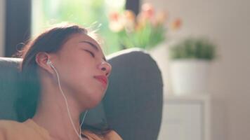 primer plano, mujer joven sentada en el sofá en la sala de estar y usar auriculares para escuchar música desde la aplicación en el teléfono inteligente y dormir en casa video