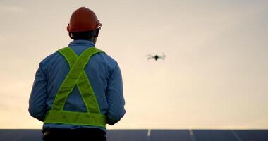 Backlit scene, Asian Young Inspector Engineer man wearing protection helmet control drone flying over row of solar panel while checking operation in solar station in evening video