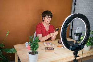 Middle-aged woman talking on cosmetics with makeup eye shadows and blush palette and brushes while recording her video. Mature female making video for her blog on cosmetics photo