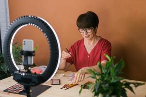 Middle-aged woman talking on cosmetics with makeup eye shadows and blush palette and brushes while recording her video. Mature female making video for her blog on cosmetics photo