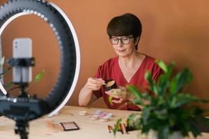 concepto de comercio electrónico y blogs de belleza y cosméticos: maquilladora profesional madura que prueba nuevos cosméticos grabando revisión de video sobre productos cosméticos en casa foto