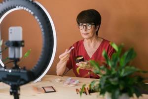 Middle-aged woman talking on cosmetics with makeup eye shadows and blush palette and brushes while recording her video. Mature female making video for her blog on cosmetics photo