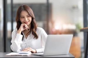 mujer joven que trabaja en una computadora portátil en la oficina. mujer de negocios asiática sentada en su lugar de trabajo en la oficina. hermosa mujer independiente que trabaja en línea en su casa. foto