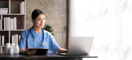 Asian doctor young beautiful woman smiling using working with a laptop computer and her writing something on paperwork or clipboard white paper at hospital desk office, Healthcare medical concept photo