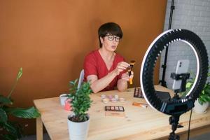 blogger de belleza femenina de mediana edad o influencer con video de grabación de productos de maquillaje en la habitación de su casa. usando lámpara de anillo y teléfono inteligente foto