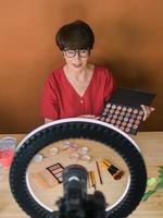 Middle-aged woman talking on cosmetics with makeup eye shadows and blush palette and brushes while recording her video. Mature female making video for her blog on cosmetics photo