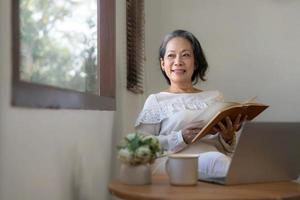 Relaxed and calm 60s retired Asian woman looking out the window, daydreaming while reading a book in her home living room. lifestyle concept photo